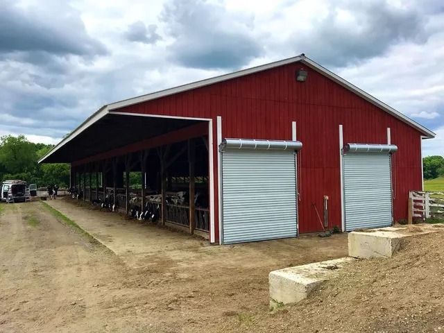 Red barn with open side shelter and silver roll-up doors on a dirt lot under a cloudy sky