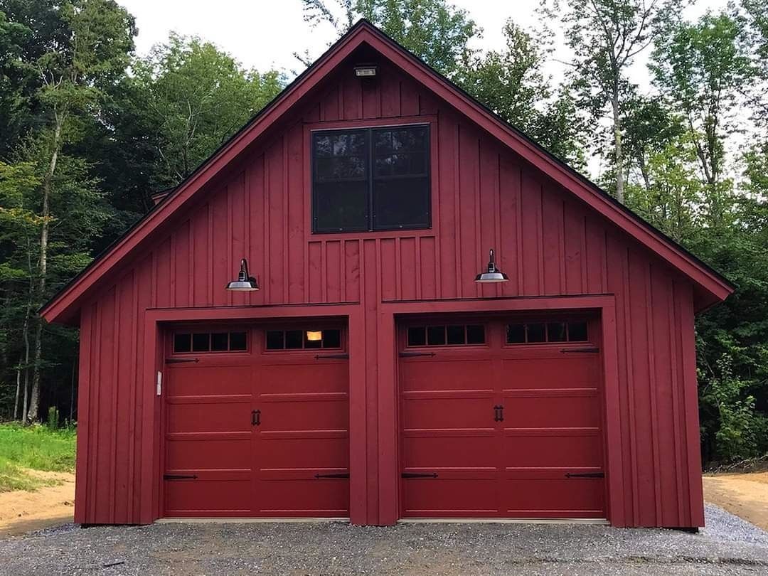 Red wooden garage with two doors and a gable roof, set among trees and gravel.