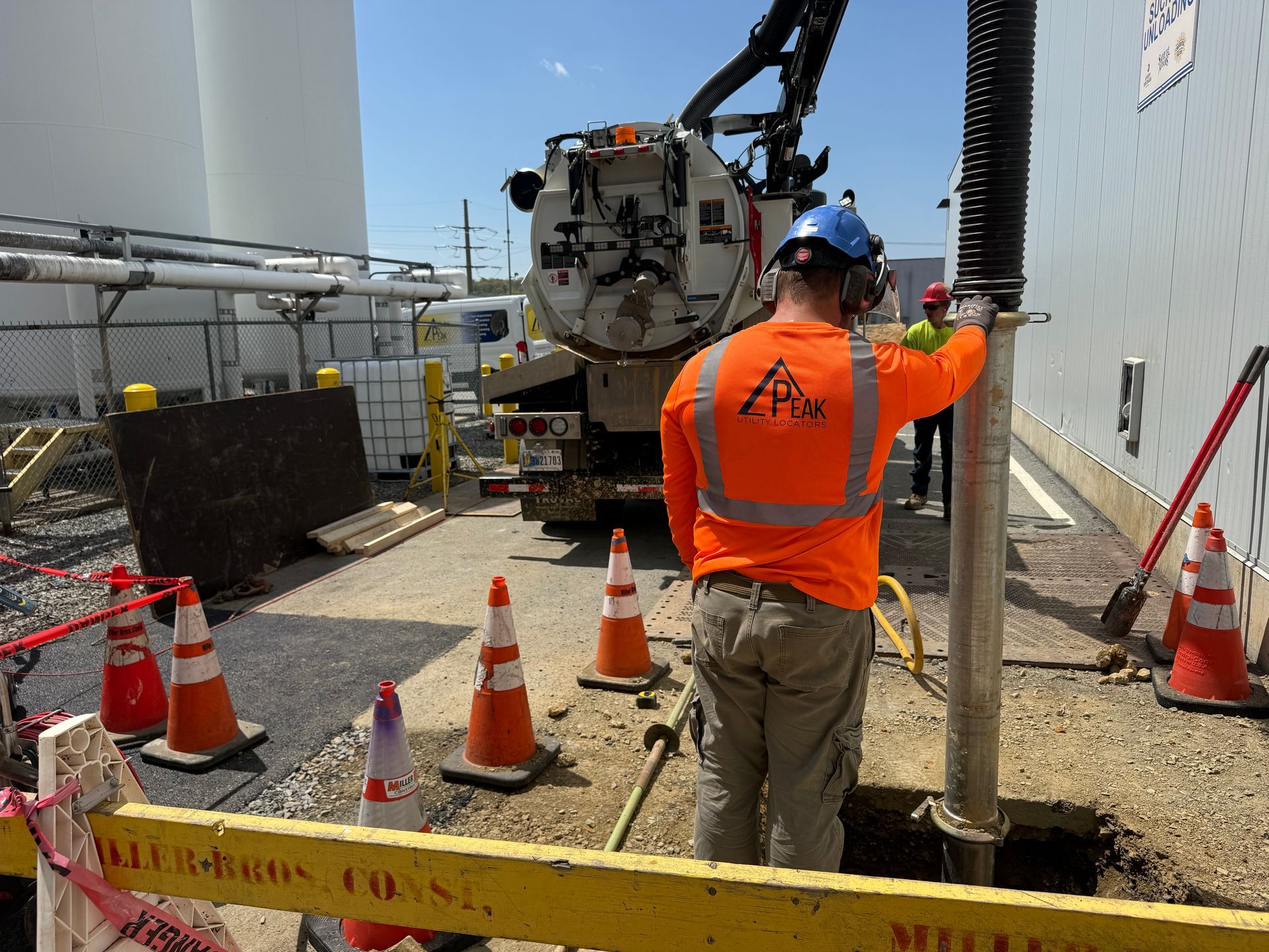 Construction worker in orange vest directs a hose beside drilling equipment and traffic cones at a worksite