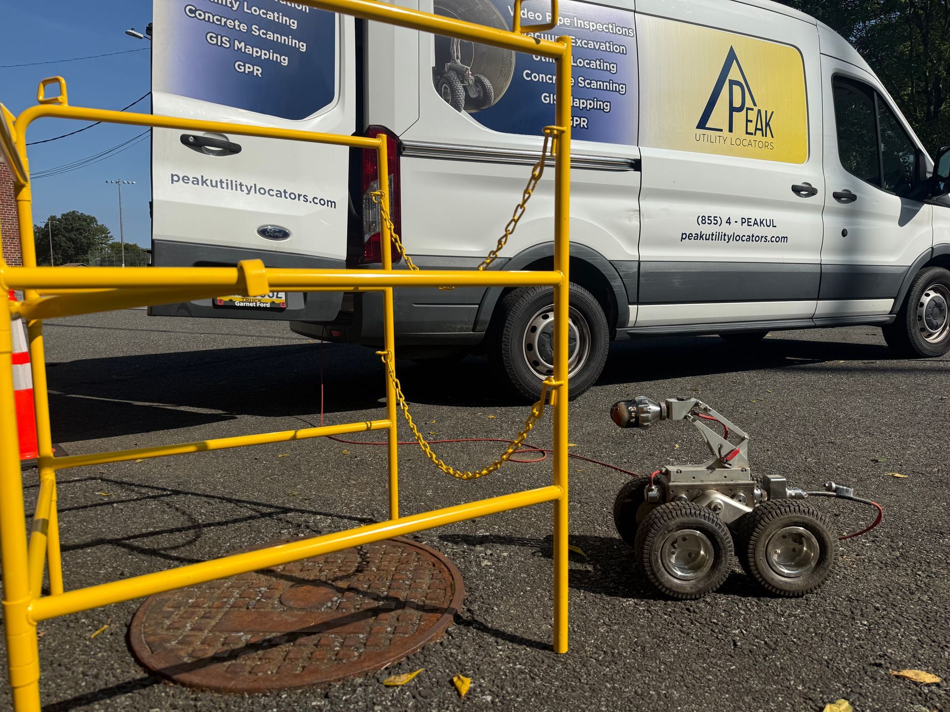 Yellow barrier in front of a white work van on asphalt, with a small wheeled machine on the ground.