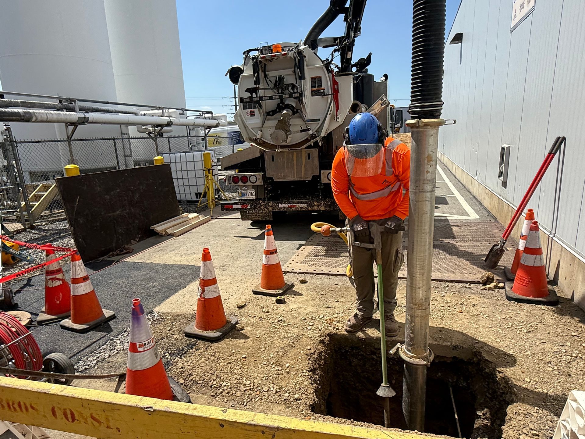 Worker in orange safety vest operating drilling equipment at a construction site, surrounded by cones and barriers