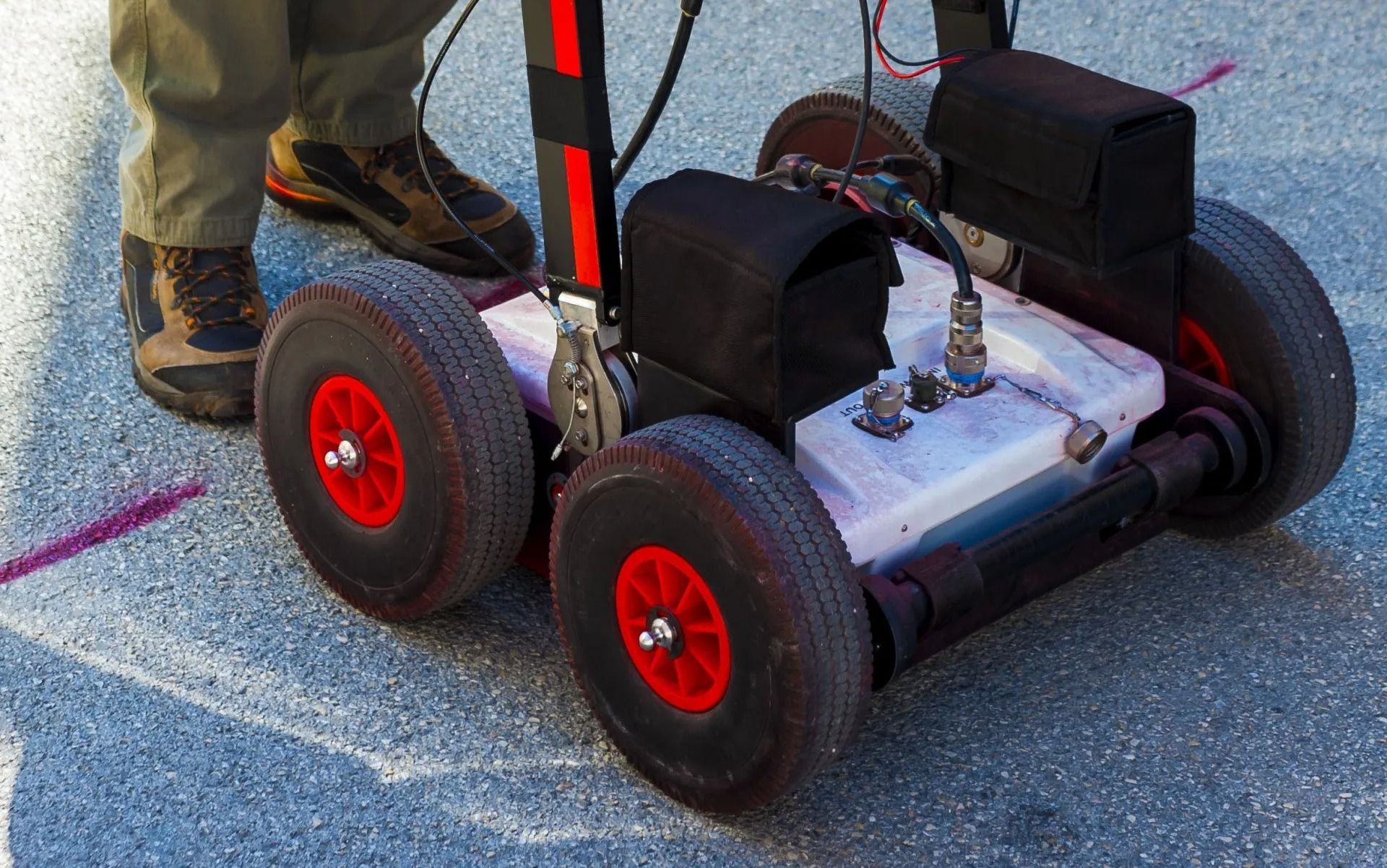 Small wheeled robot with red wheels on pavement beside a person’s boots and legs