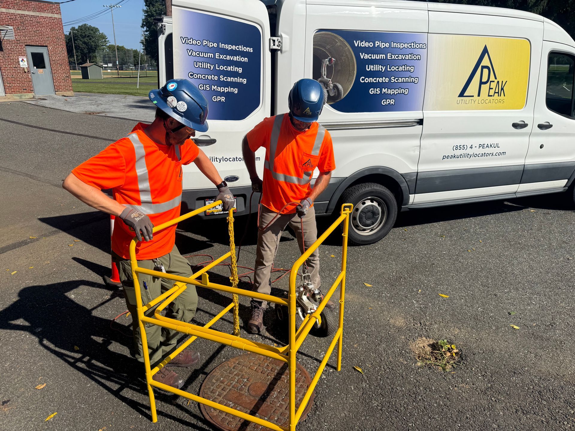 Two workers in orange safety vests inspect a yellow metal platform beside a utility van.