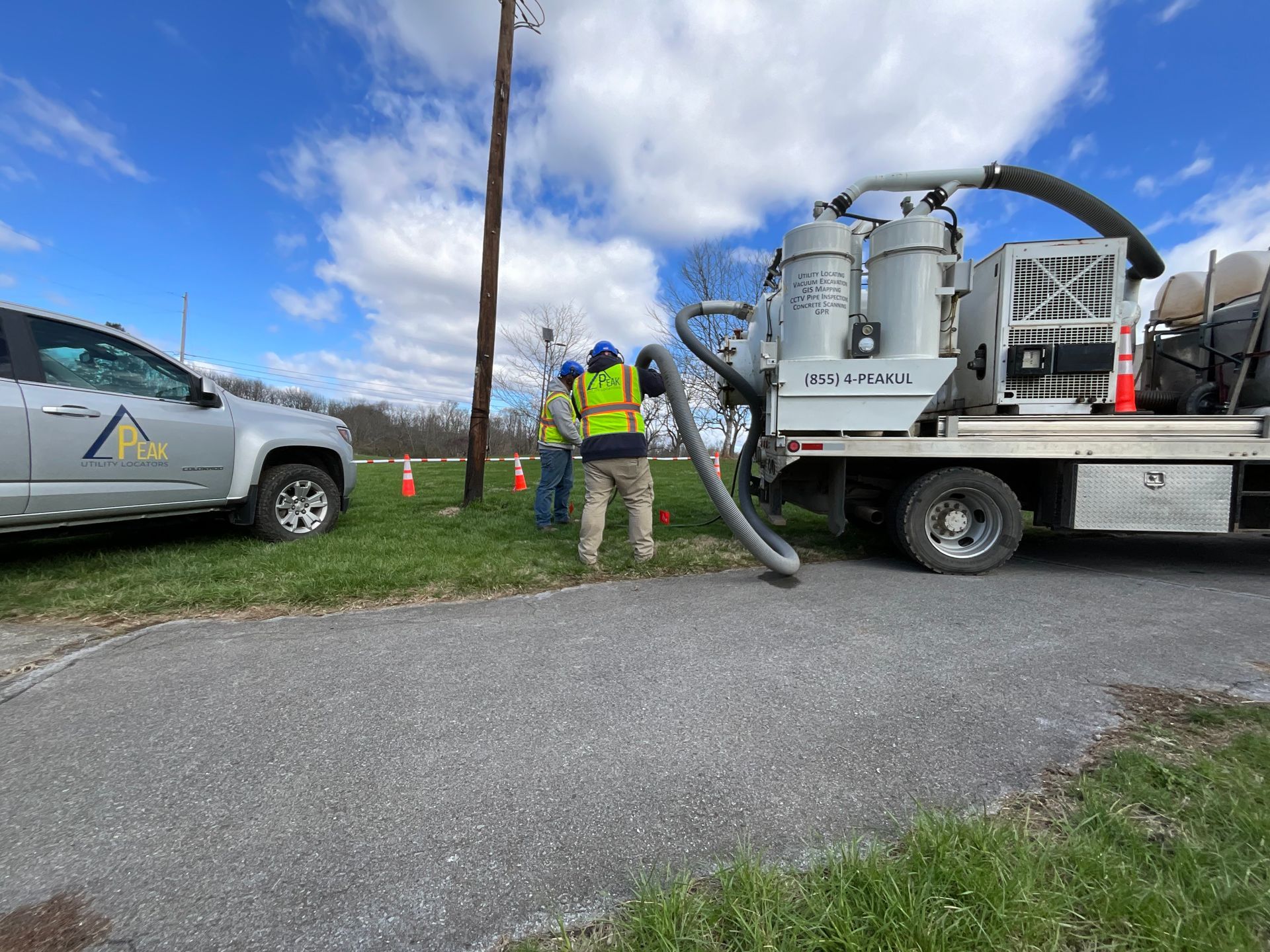 Utility workers inspecting a large hose on a truck beside a roadside under a cloudy sky