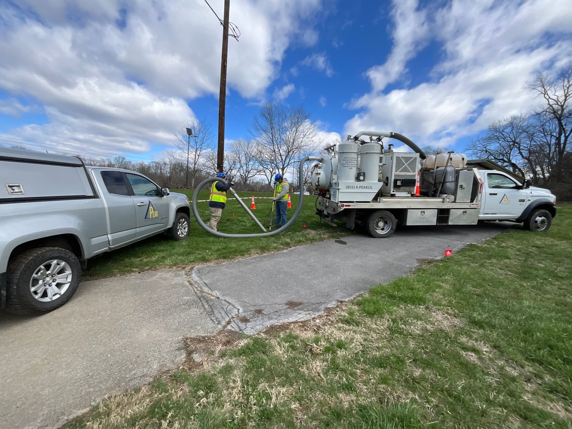 Utility trucks working beside a roadside utility pole, with hoses and equipment on the grass under a cloudy sky