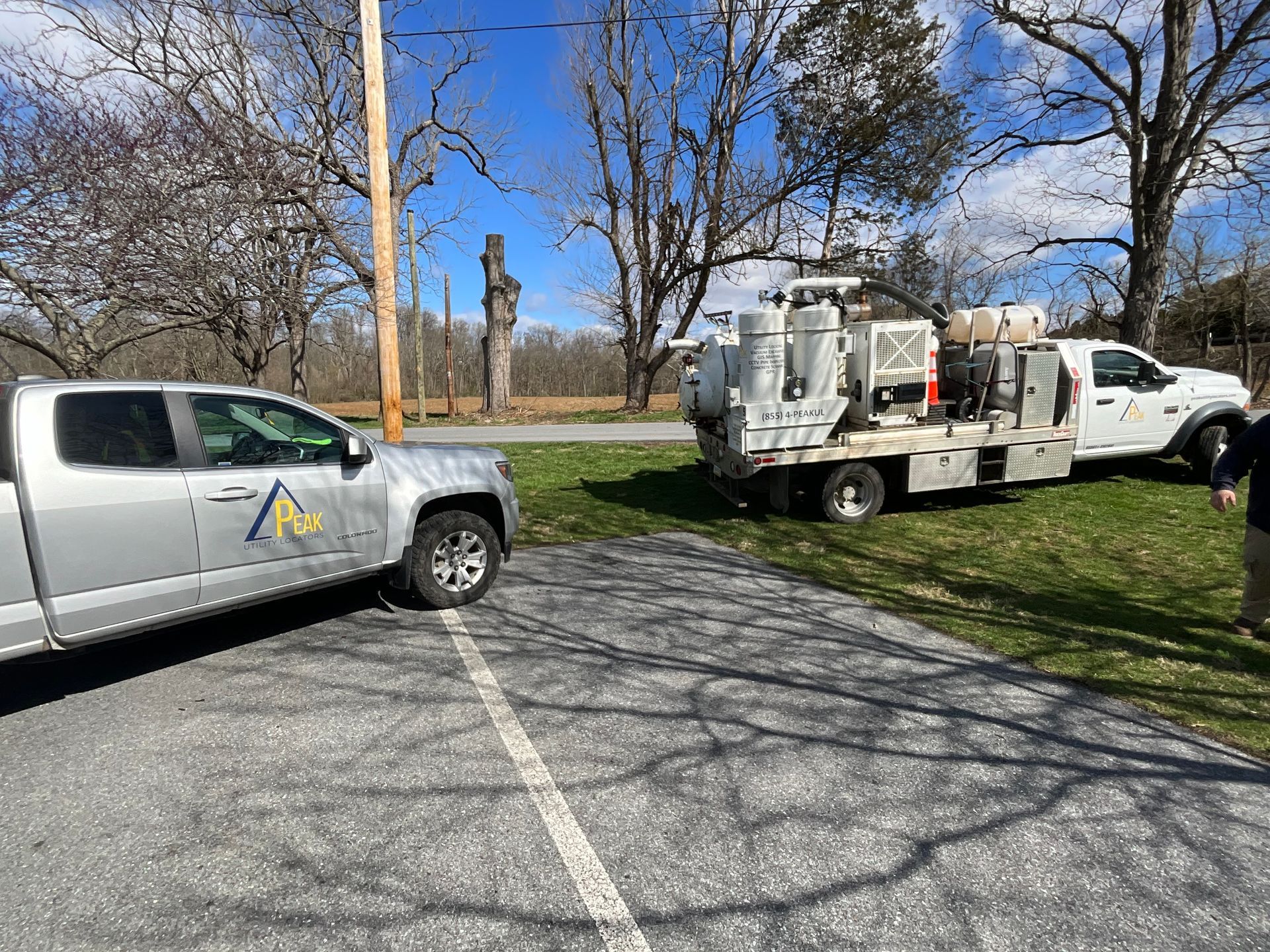 Utility trucks parked by a grassy roadside under leafless trees on a sunny day
