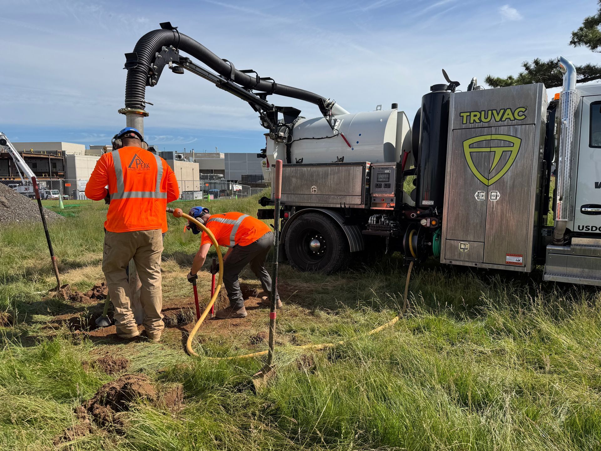 Utility workers in orange vests use a truck-mounted drill in a grassy roadside work site.