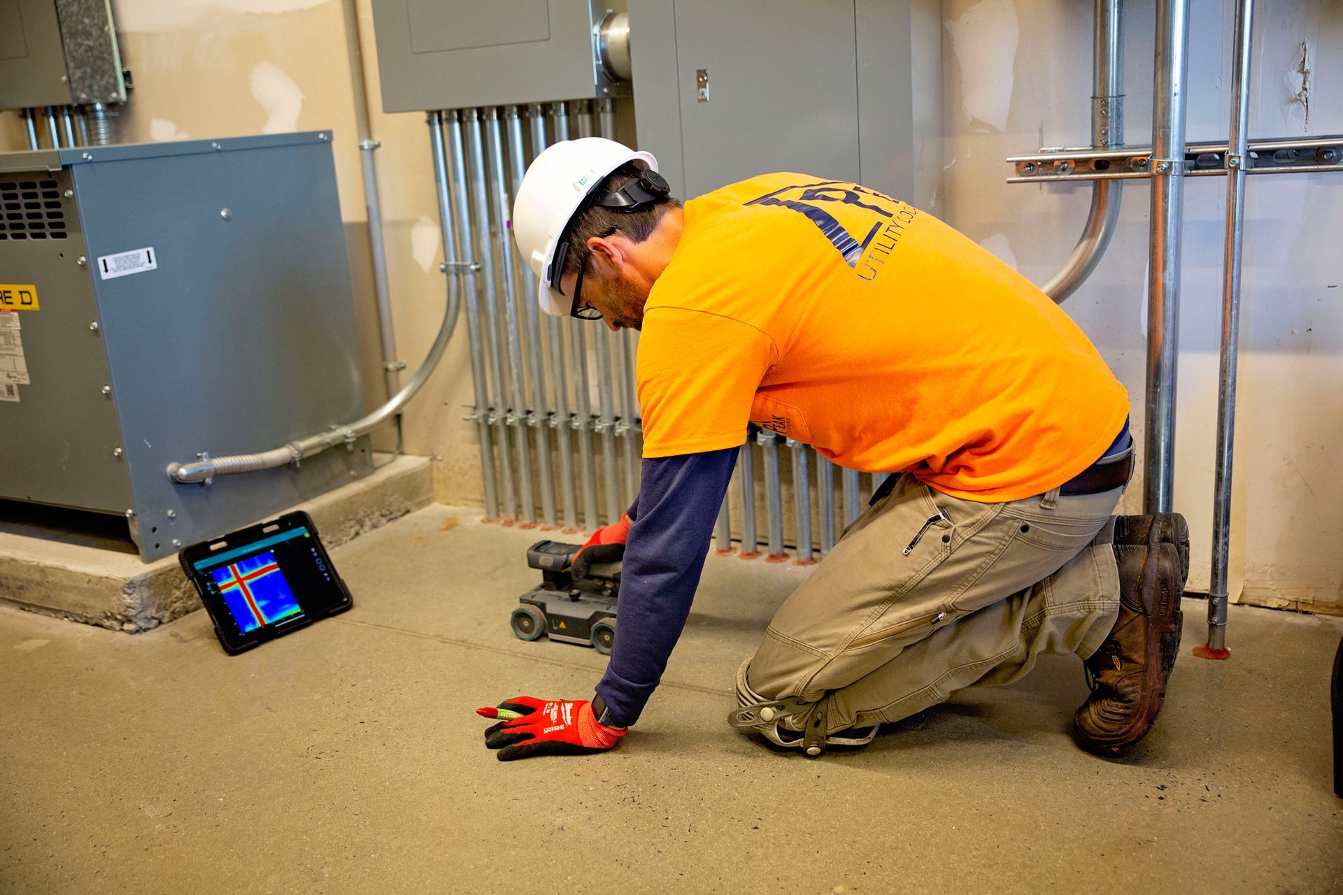 Technician kneeling on floor beside a tablet, inspecting equipment in a utility room