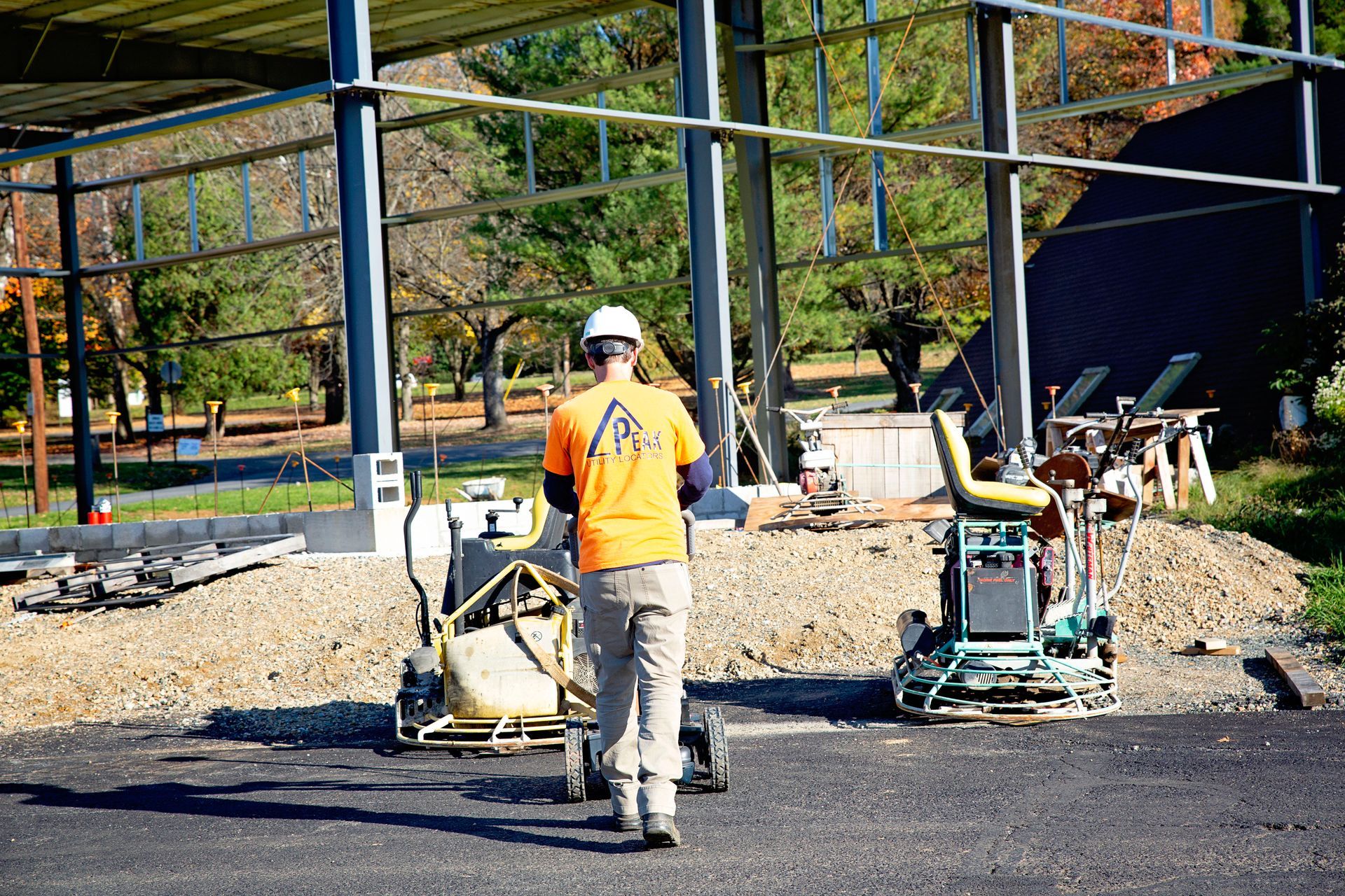 Worker using a paving machine on a gravel lot beside a steel-frame building under construction