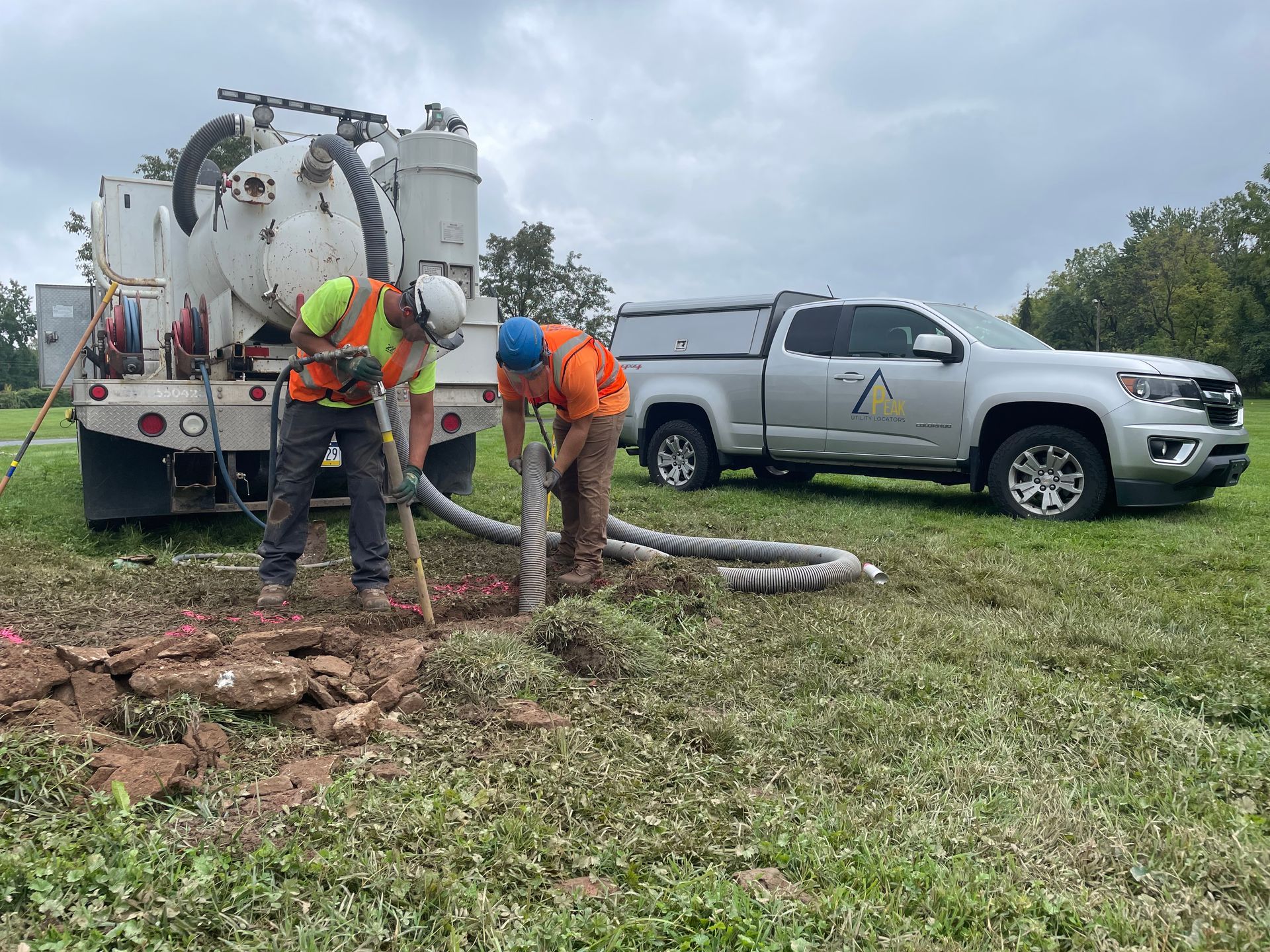 Utility workers beside a tanker truck and pickup, handling hoses in a grassy field