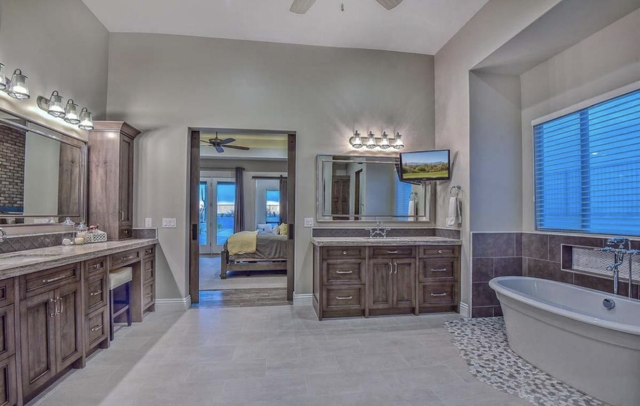 A bathroom with two sinks, wooden cabinets, a tub, and a television