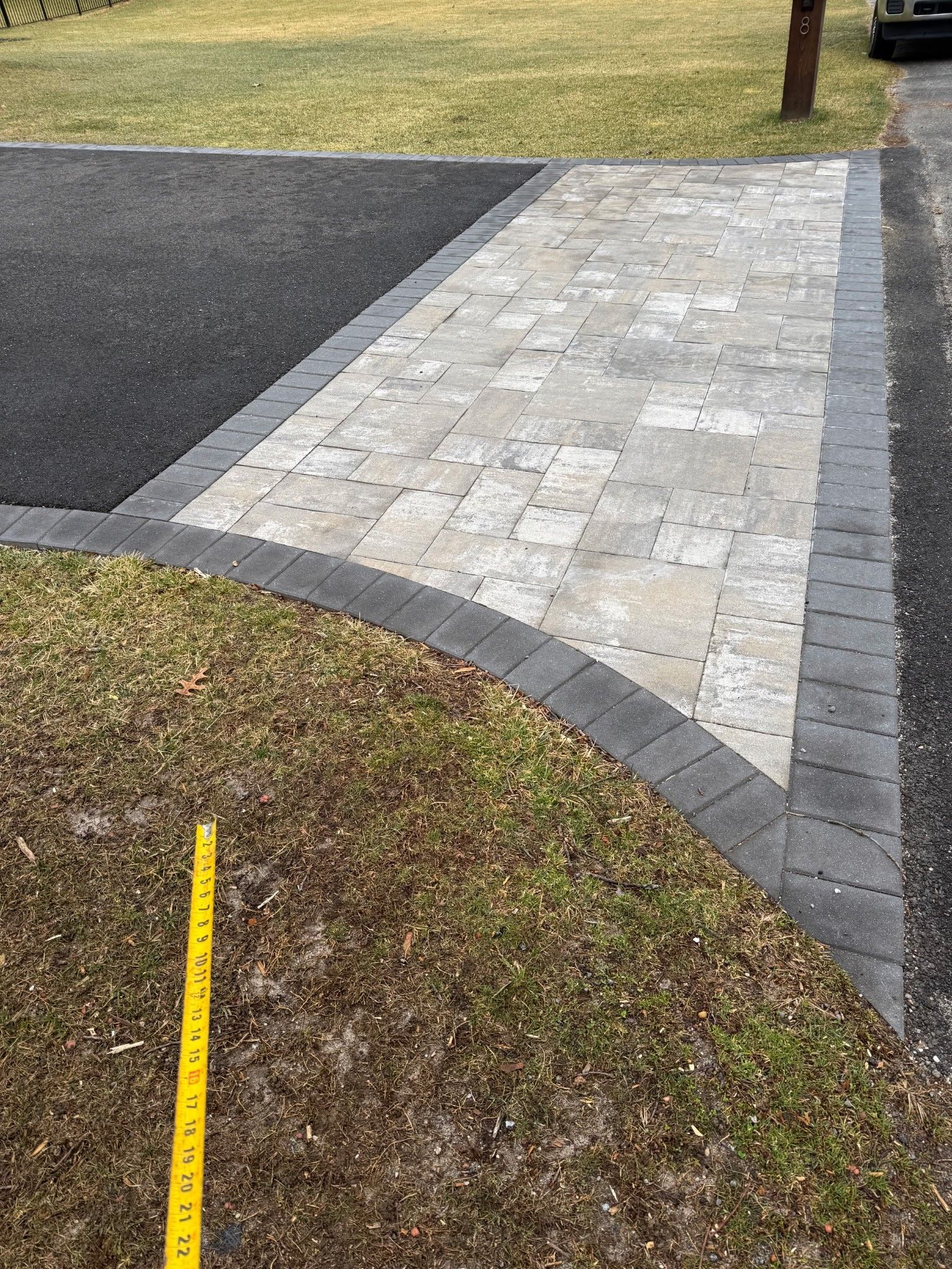 Driveway and walkway with light stone pavers, bordered by dark gray bricks and grass.