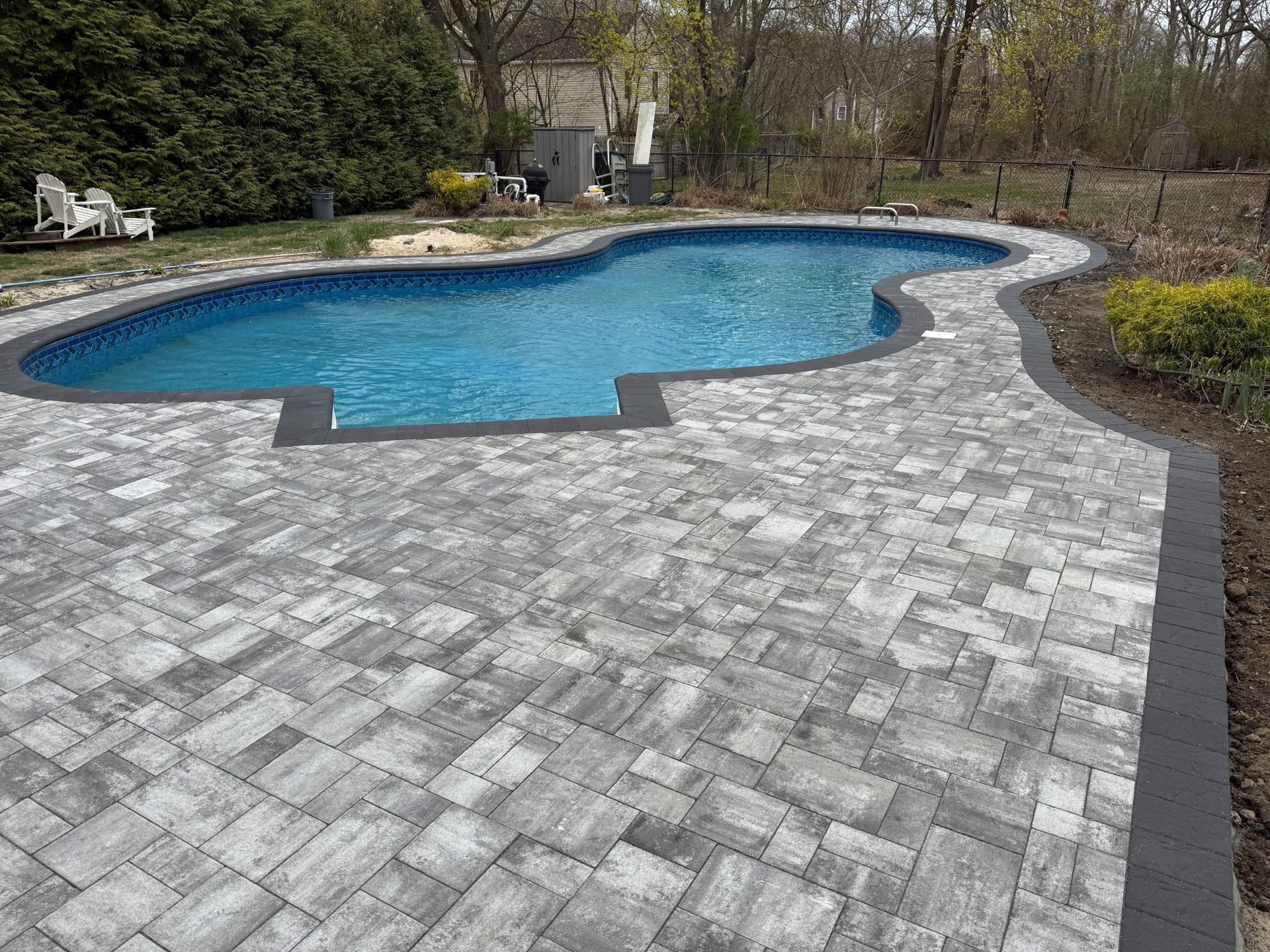 Curved backyard pool with blue water and gray stone paver patio, bordered by landscaping.
