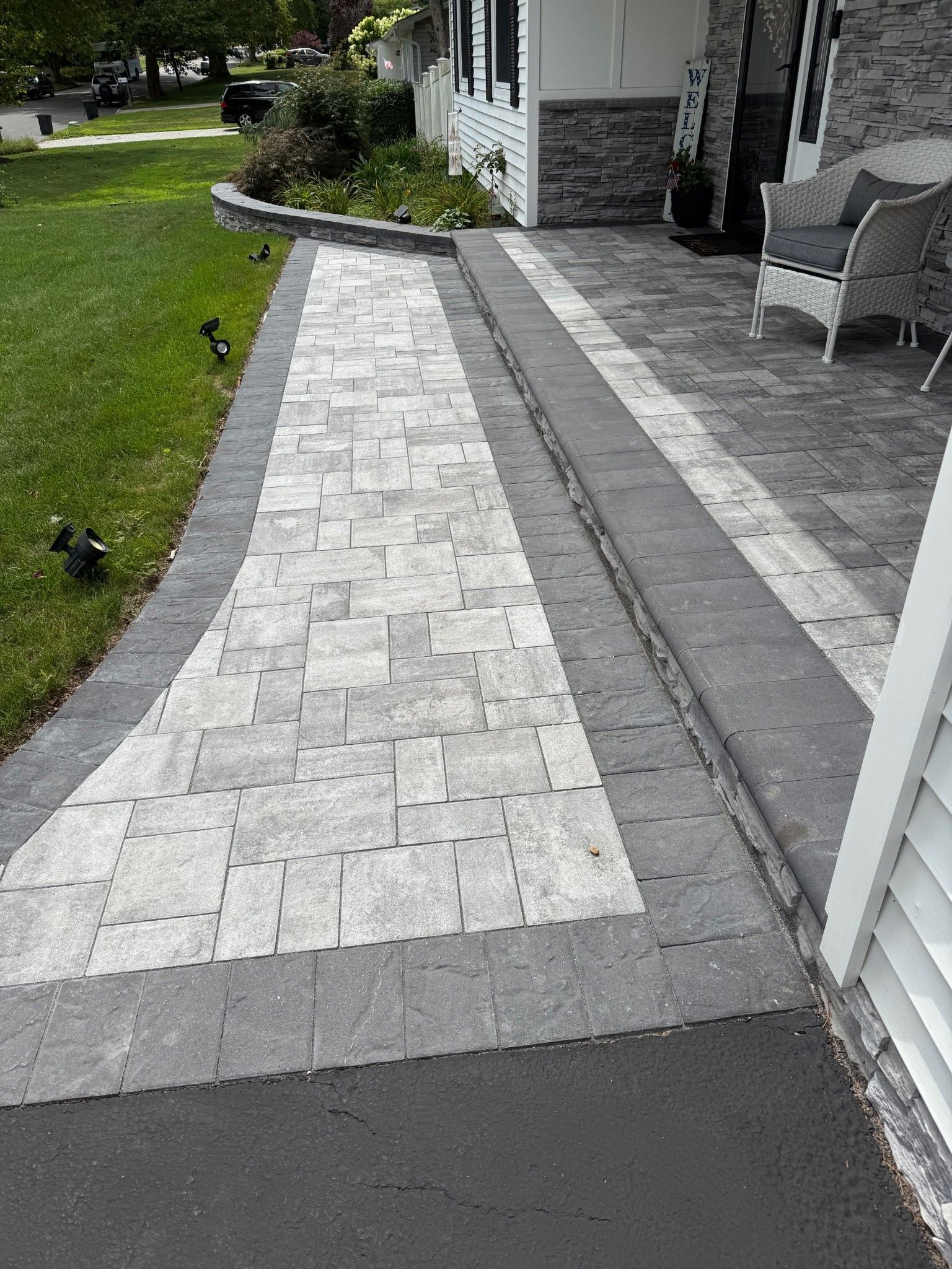 Brick walkway beside a house with a gray paver patio and green lawn.