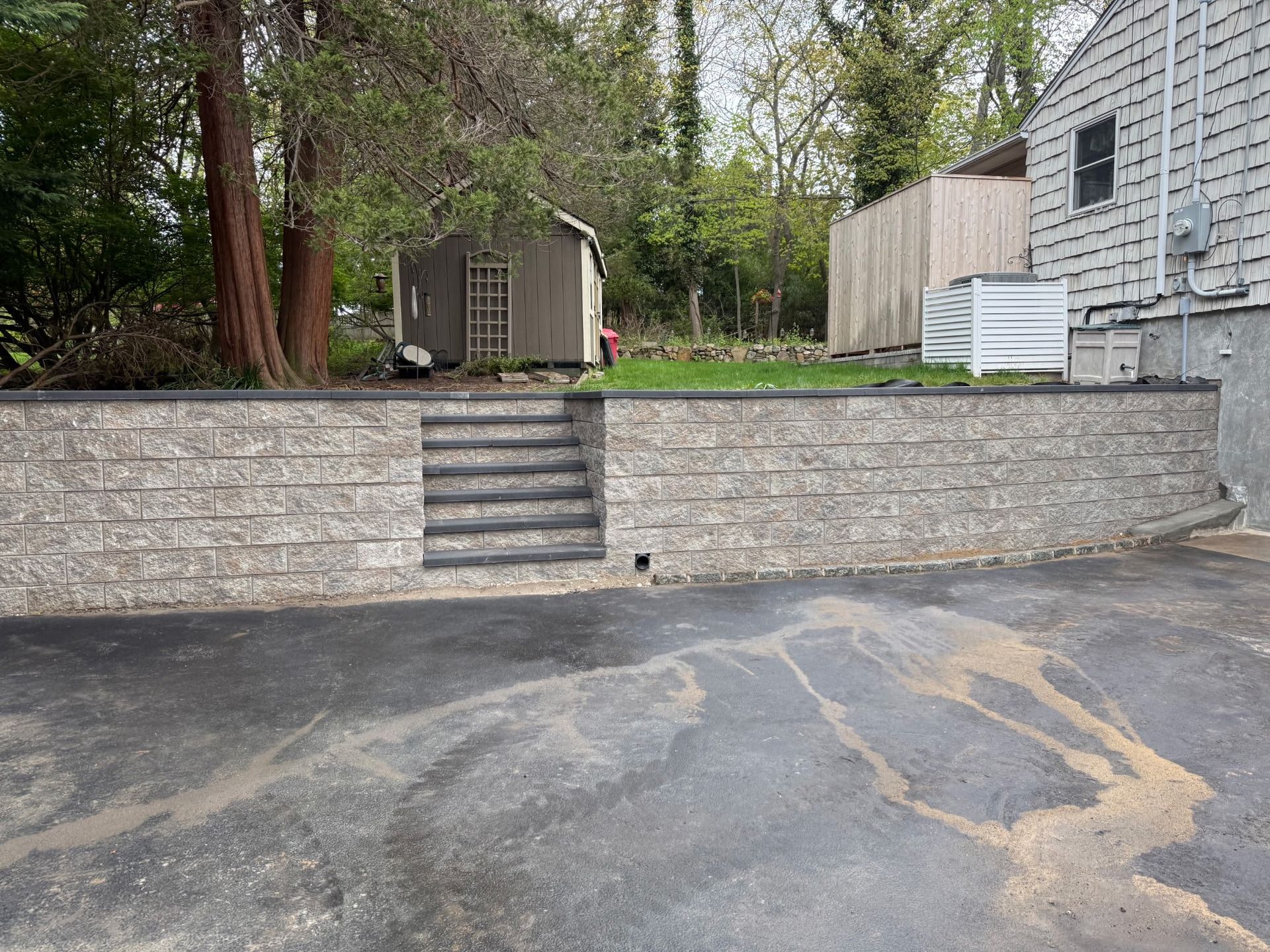 Backyard with stone retaining wall, central stairs, shed, and a cracked asphalt driveway.