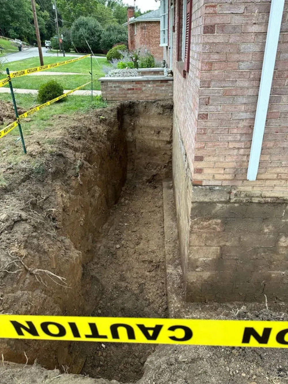 Trench dug next to a brick building. Yellow caution tape. Soil, grass, and a street are in the background.