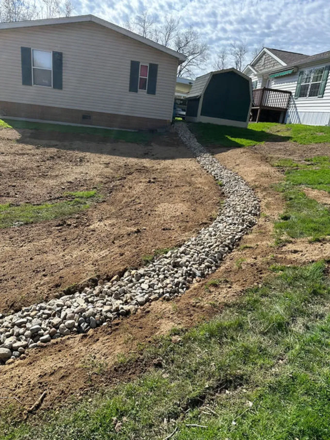 A gravel-lined drainage channel curves through a grassy yard, near a light-colored house and shed.
