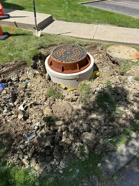 Open manhole in a dirt pit on a grassy area near a sidewalk and road. Orange cones and a shovel are nearby.