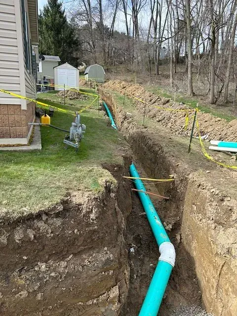Trench dug for installing blue PVC pipes in a grassy backyard. Yellow caution tape is present.