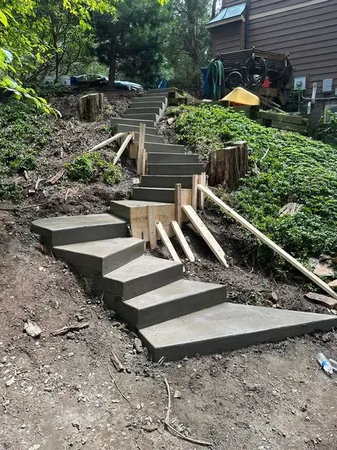 Concrete steps under construction on a hillside, wooden supports, and surrounding greenery.