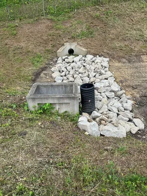 Concrete culvert draining into a rock-lined channel with a corrugated plastic pipe and a concrete basin.