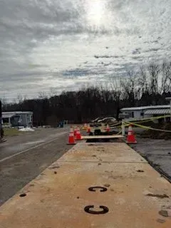 Roadway with open hole, marked with orange cones and caution tape. Cloudy sky, trees in the background.