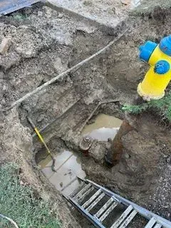 Excavation reveals water pipes and a ladder, next to a yellow and blue fire hydrant.