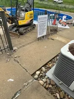 Mini excavator next to a pool under construction, concrete sidewalk has gaps filled with river rock.