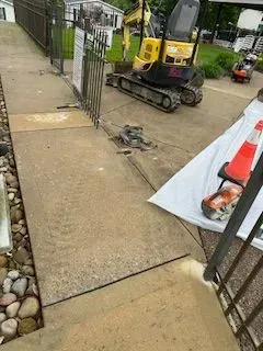 Sidewalk repair: Mini excavator and tools on concrete; orange safety cone visible.