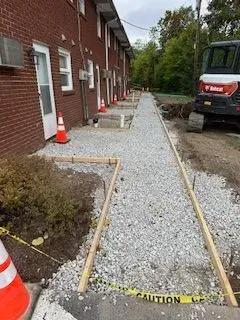 Sidewalk construction at apartment building, gravel base in wooden frame, Bobcat excavator in the background.