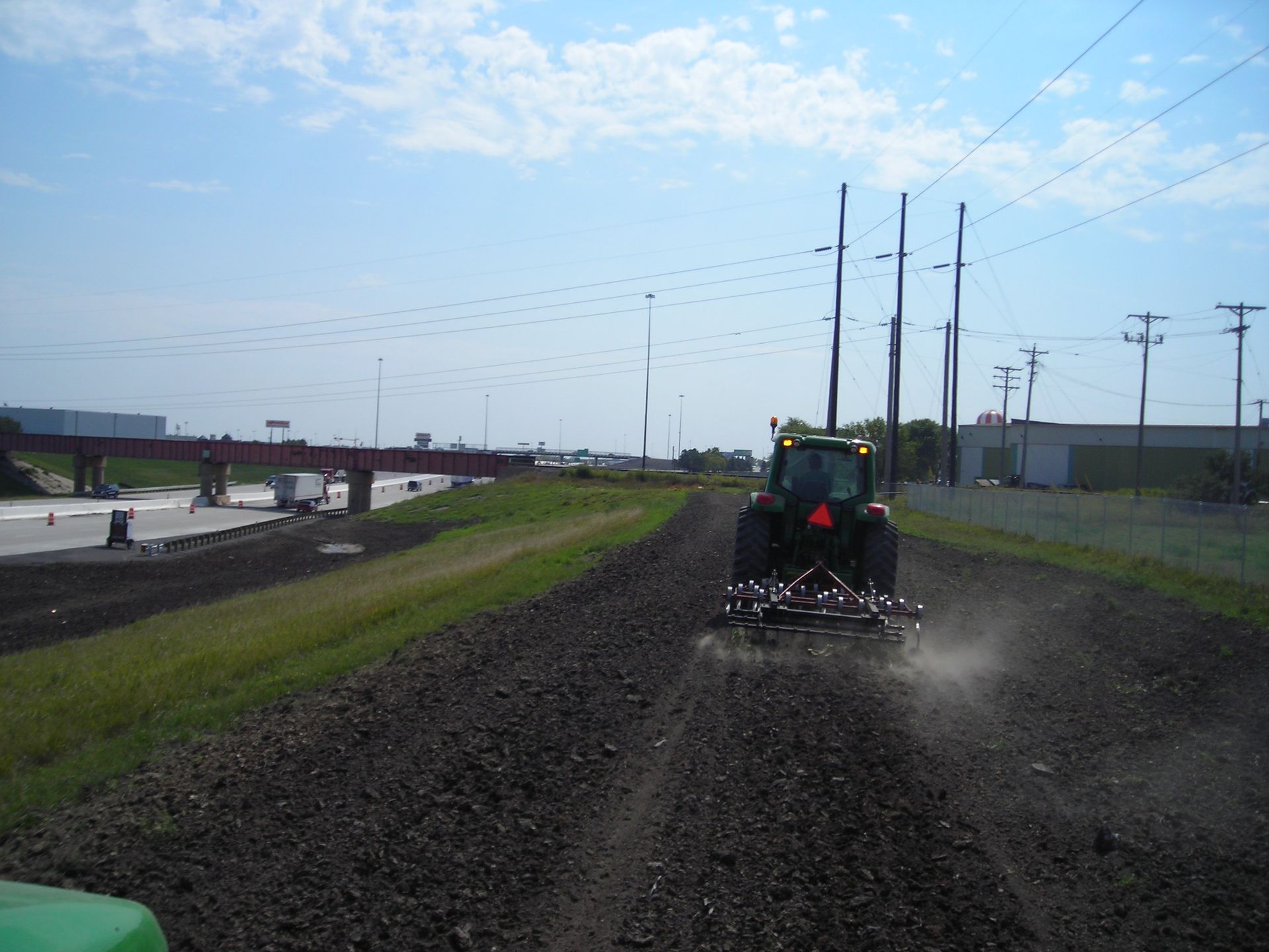 A tractor is plowing a dirt road with a bridge in the background