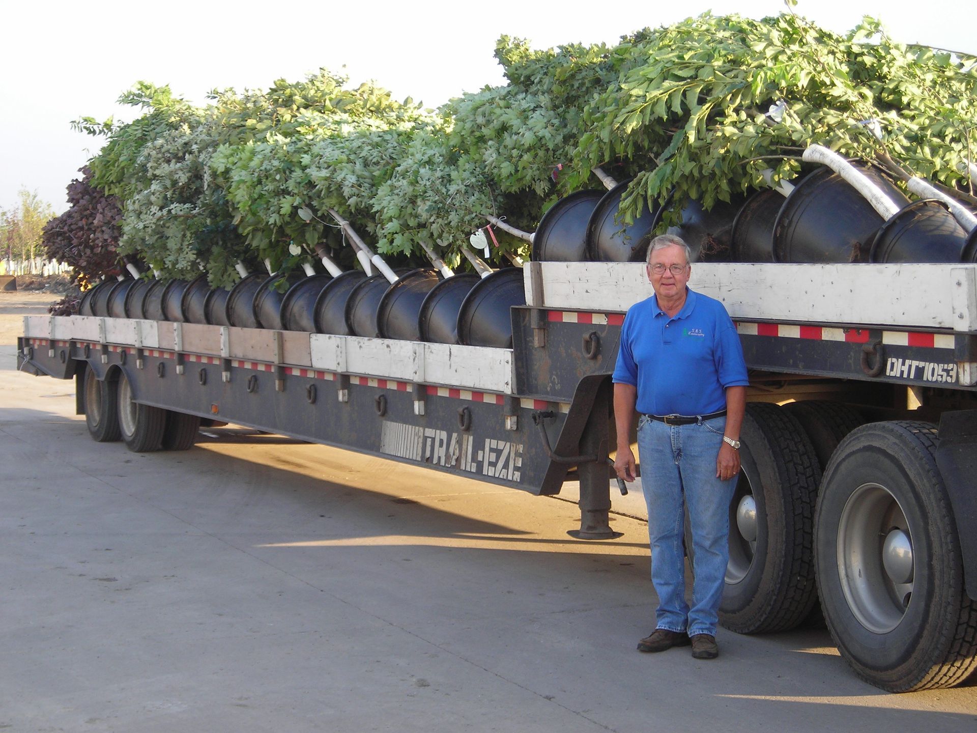 A man is standing in front of a semi truck with trees on it