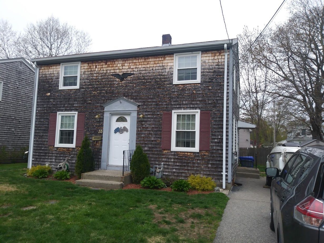 Two-story house with weathered wood siding, white door, windows, and red shutters; front yard with grass and bushes.