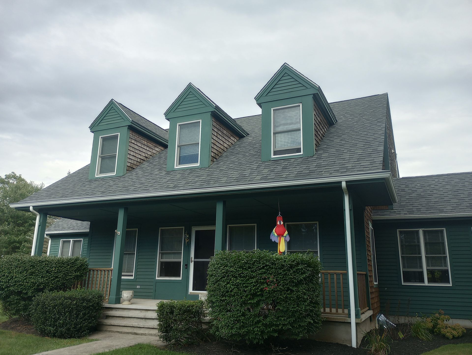 Green house with three dormers, front porch, gray roof, and bushes.