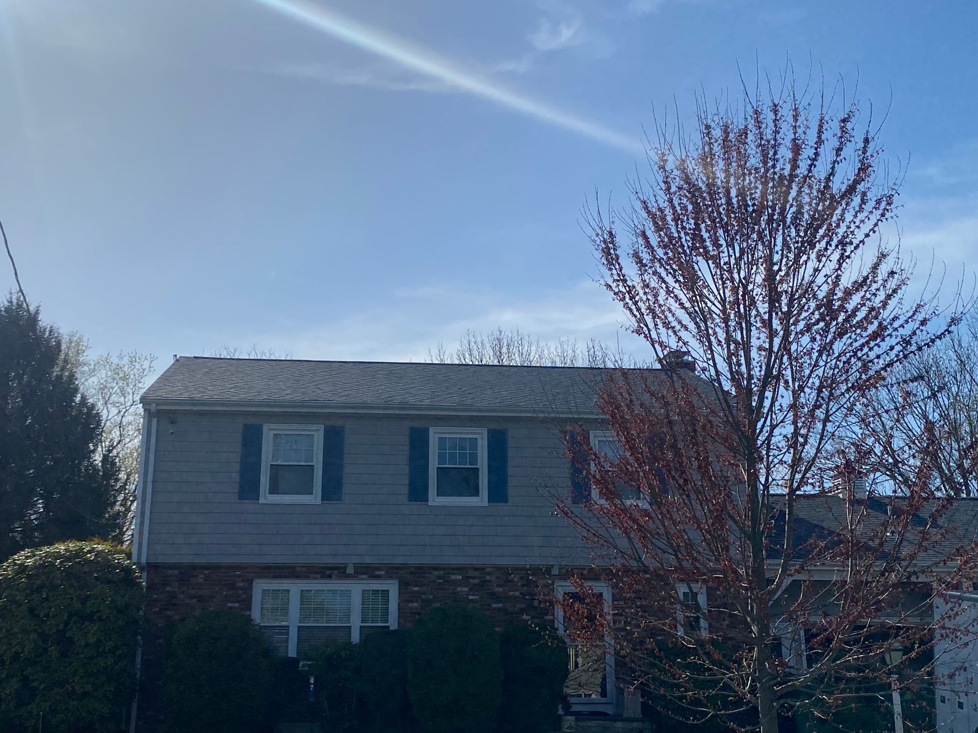 Gray house with dark shutters, red door, and a tree with red buds under a blue sky.
