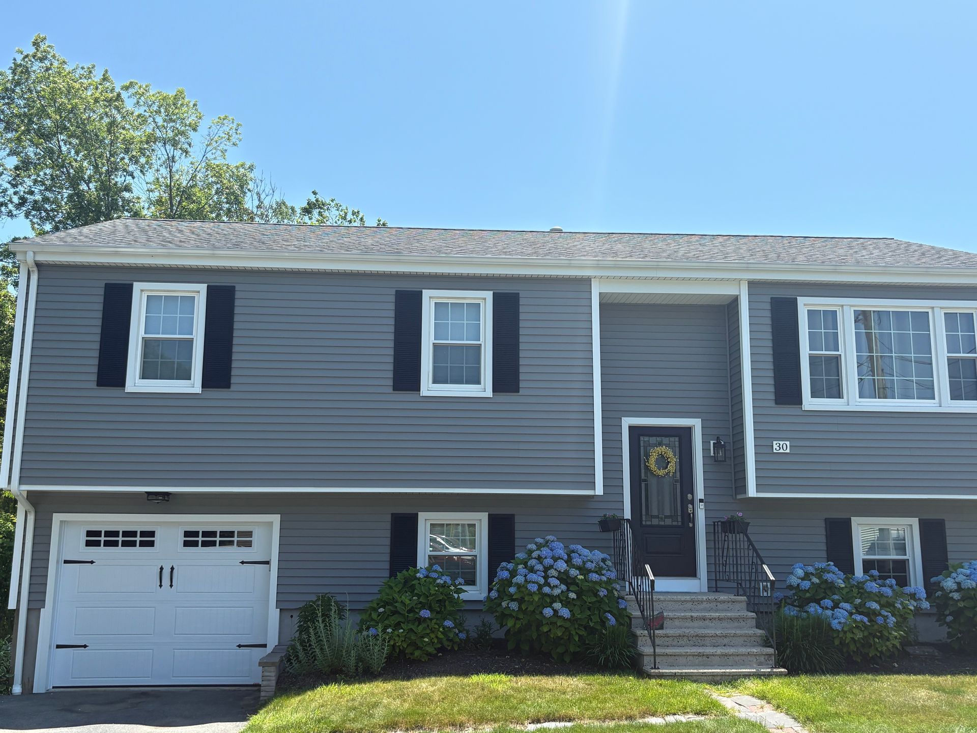 Gray house with white garage door, black shutters, and blue hydrangeas under a clear sky.