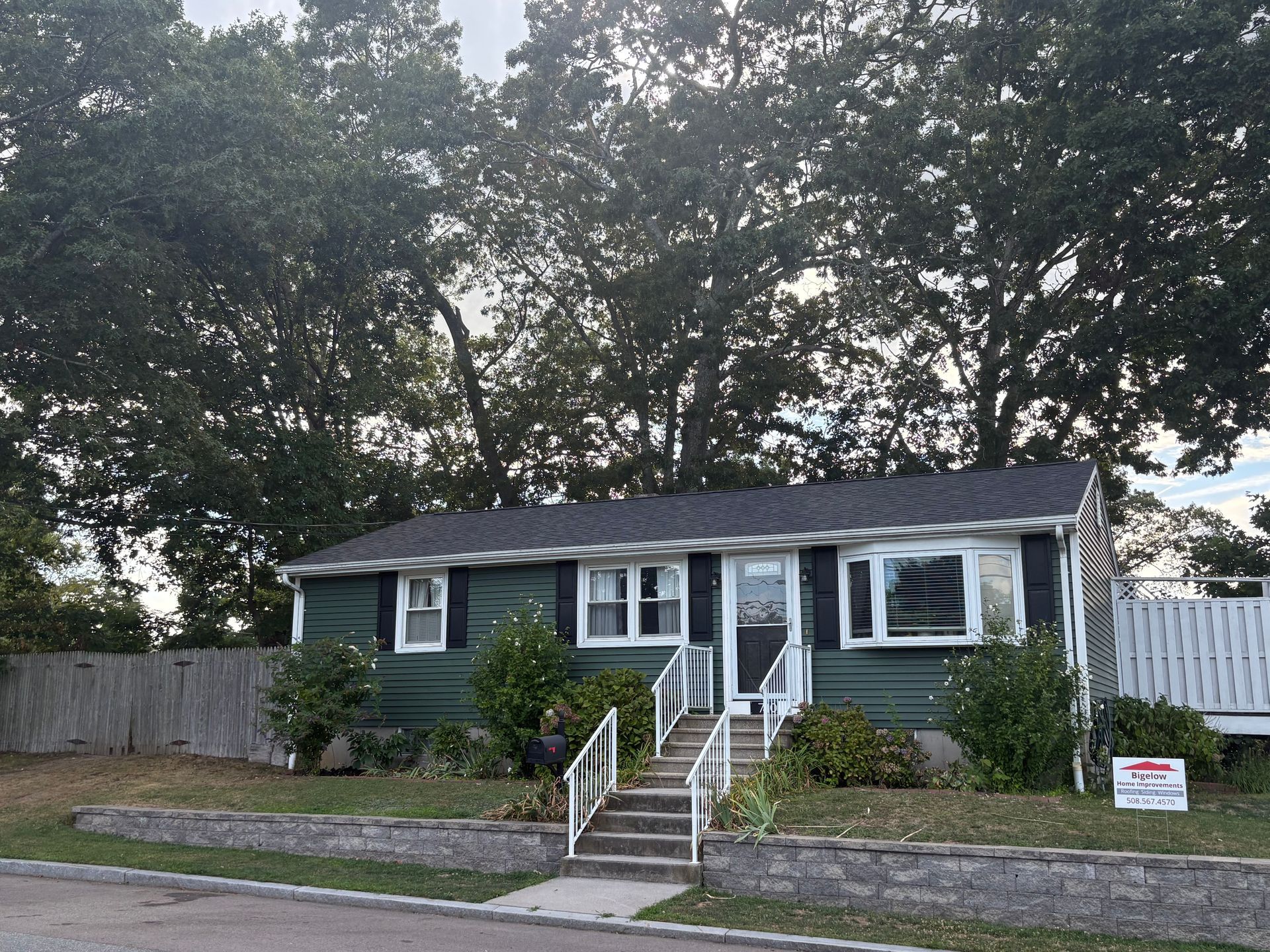 Green bungalow house with white trim, a short stairway, and a fence, with trees in the background.