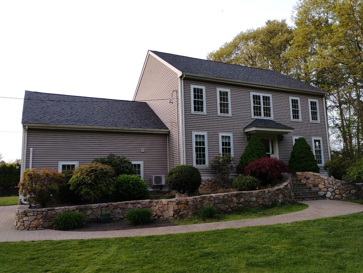 Two-story house with gray siding, dark roof, and stone retaining wall with landscaping.