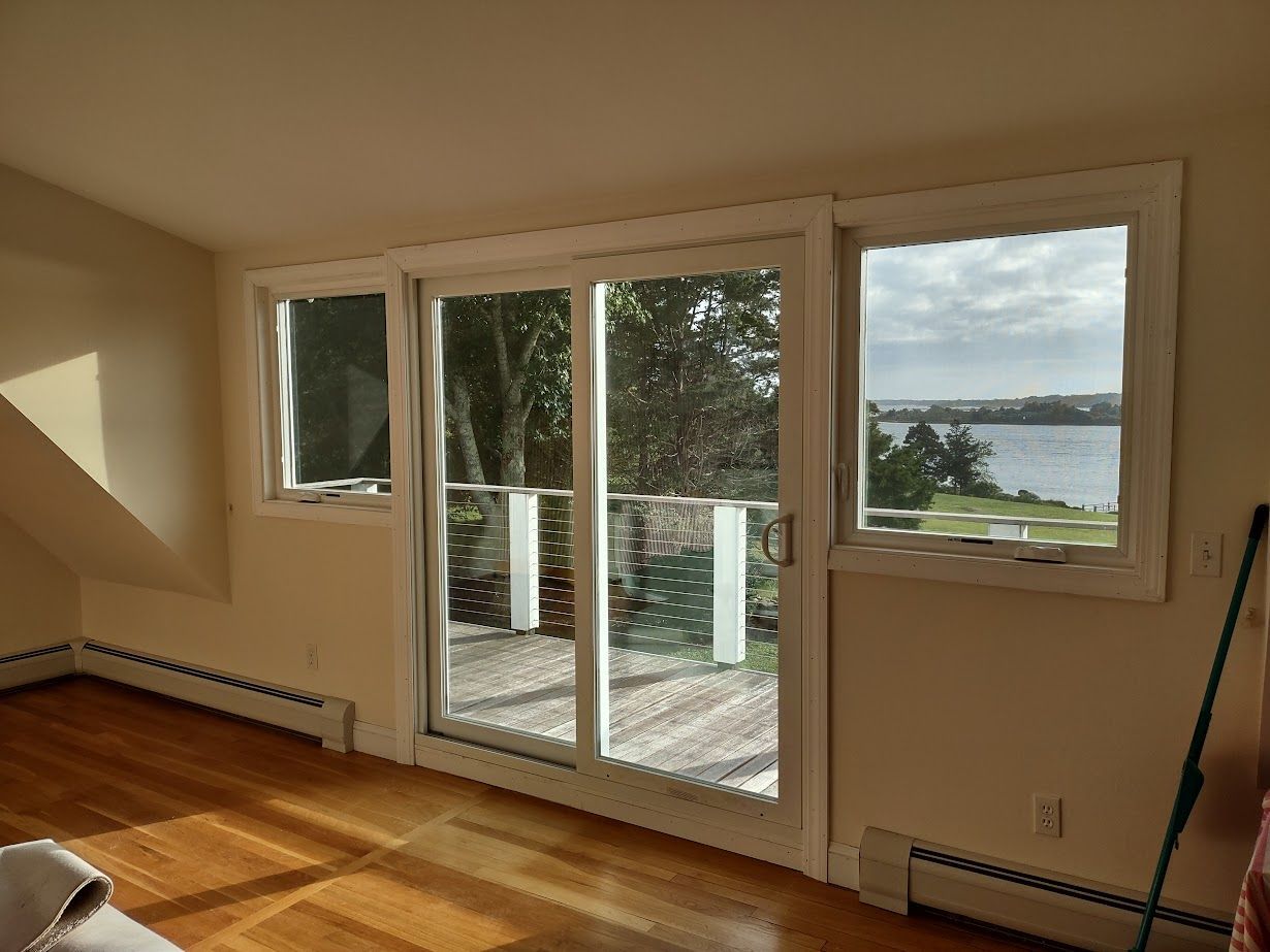 Sunlit interior with sliding glass door and two windows overlooking a body of water and trees.