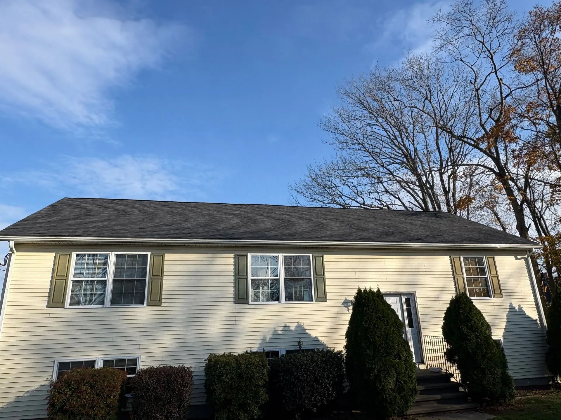 Beige house with a dark roof and green shutters against a partly cloudy blue sky.