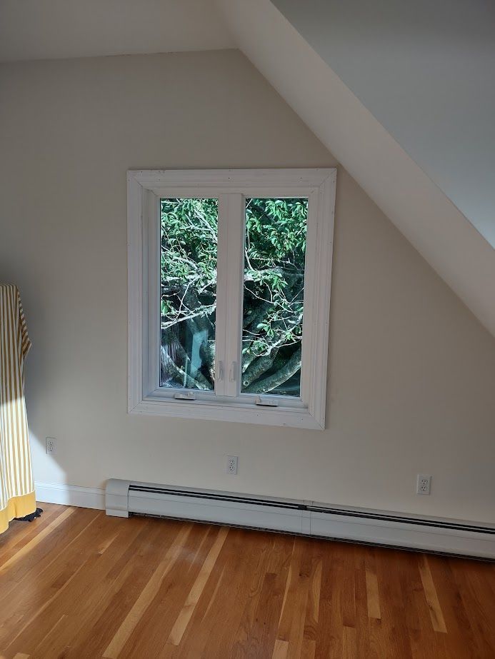 Window in a room with hardwood floors and a radiator, framed by white molding, with a view of greenery.
