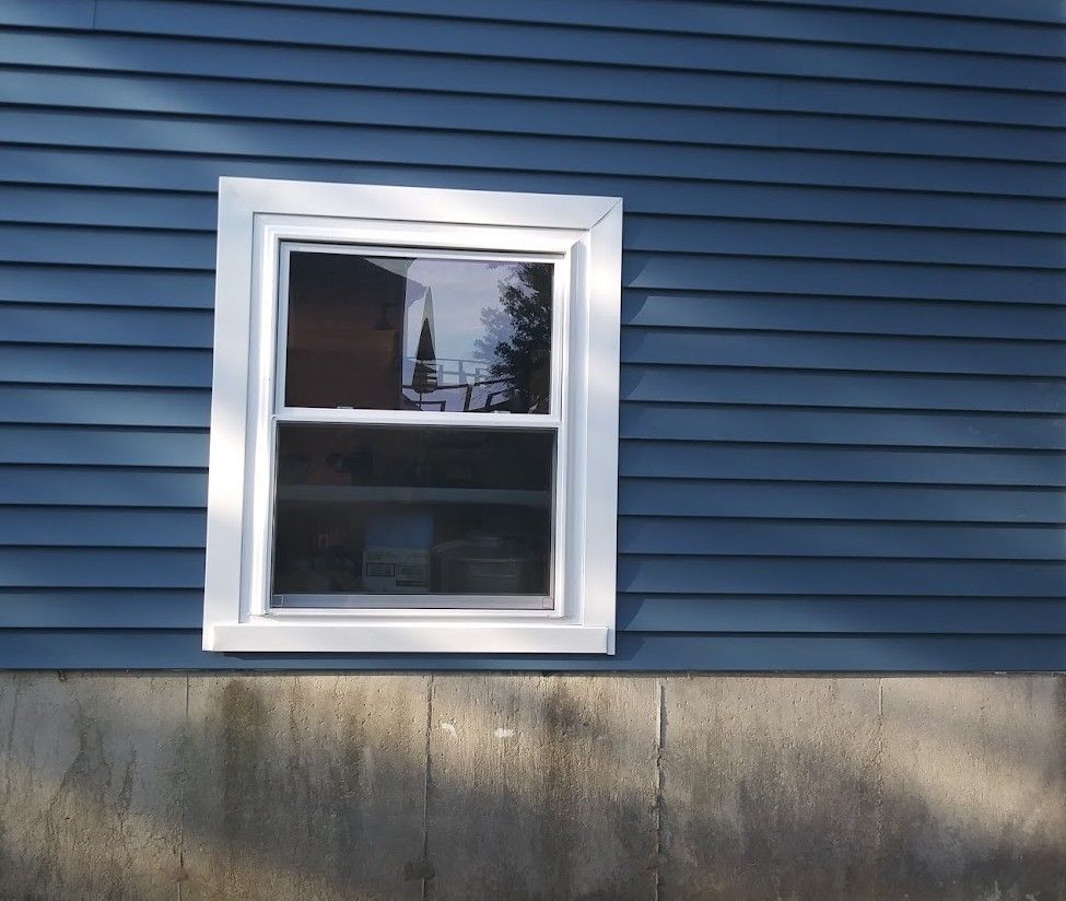 White-framed window on a blue-sided house with a concrete foundation.
