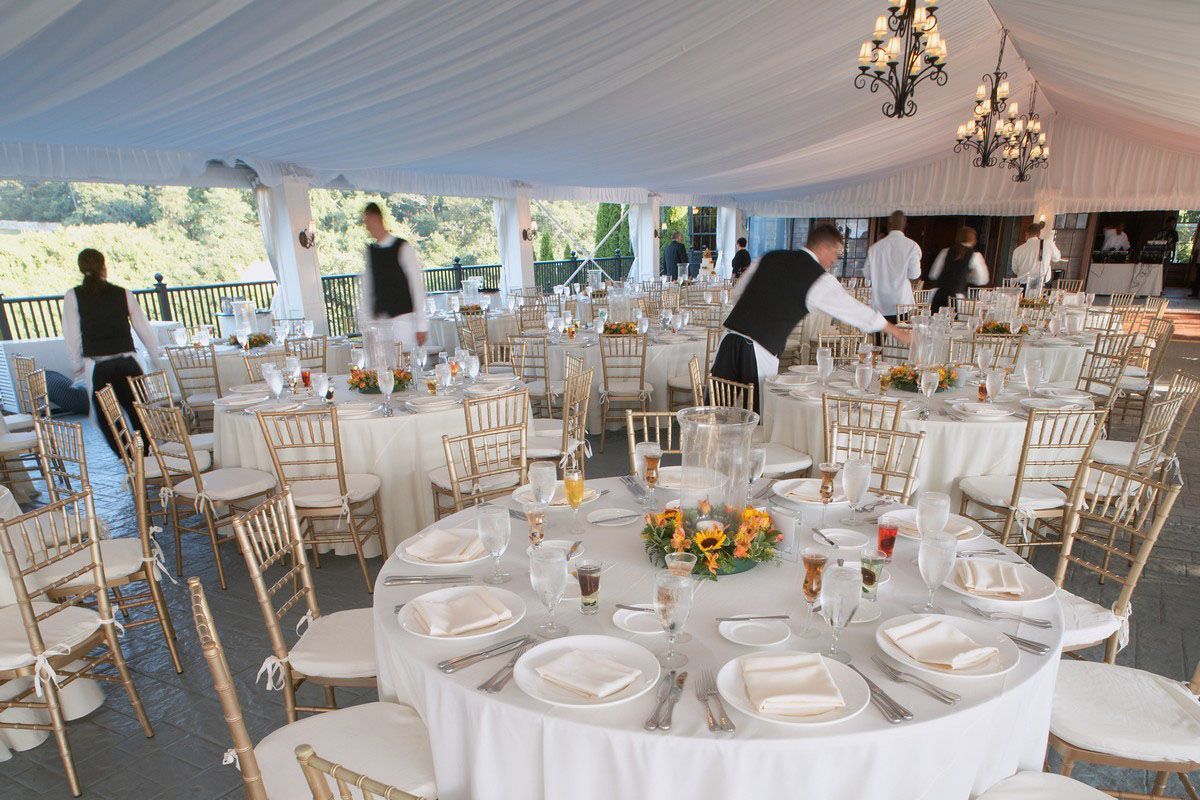 Elegant white-tented event hall with round tables, gold chairs, chandeliers, and staff setting up centerpieces