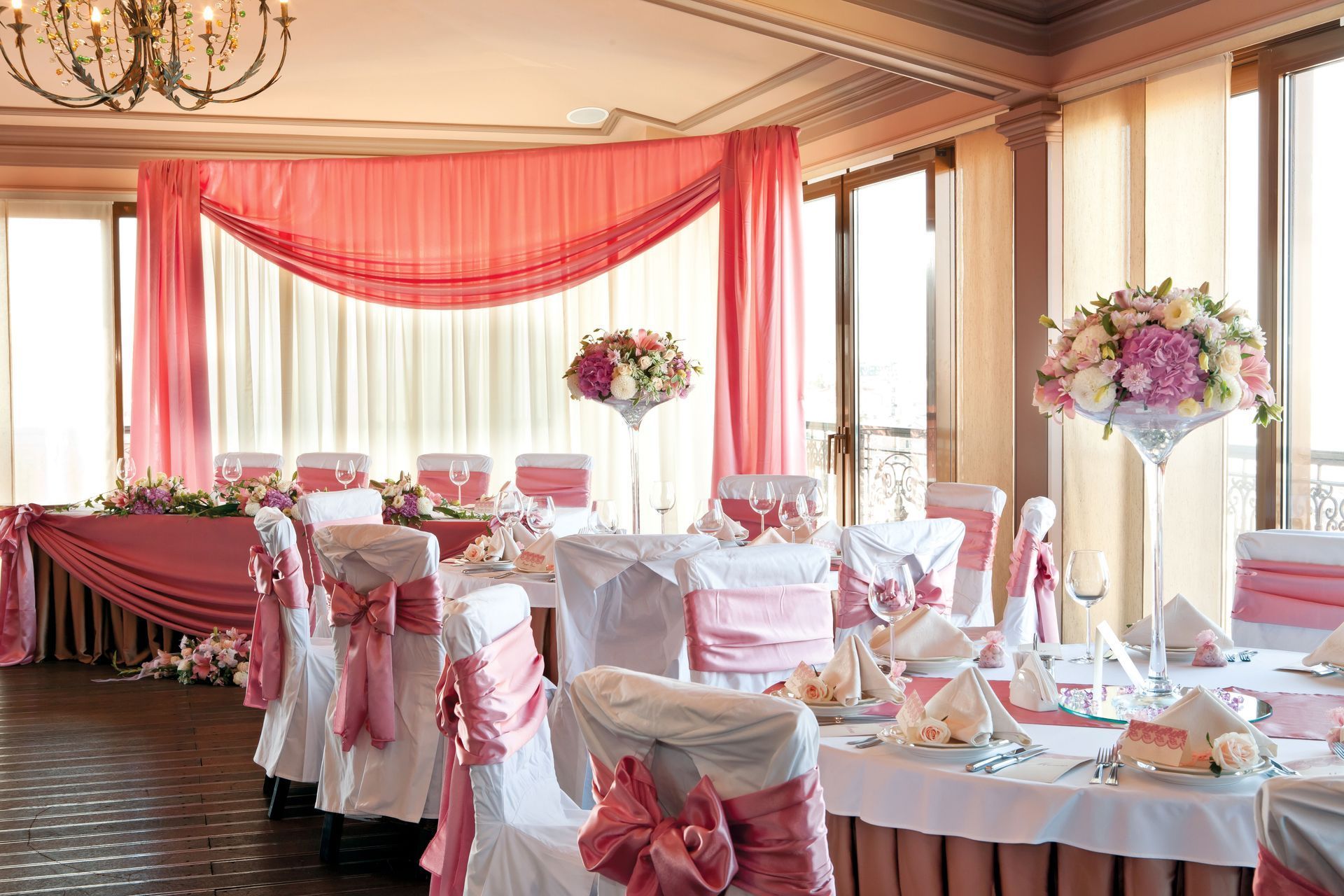Elegant banquet hall with pink drapes, white-covered chairs, round tables, and floral centerpieces
