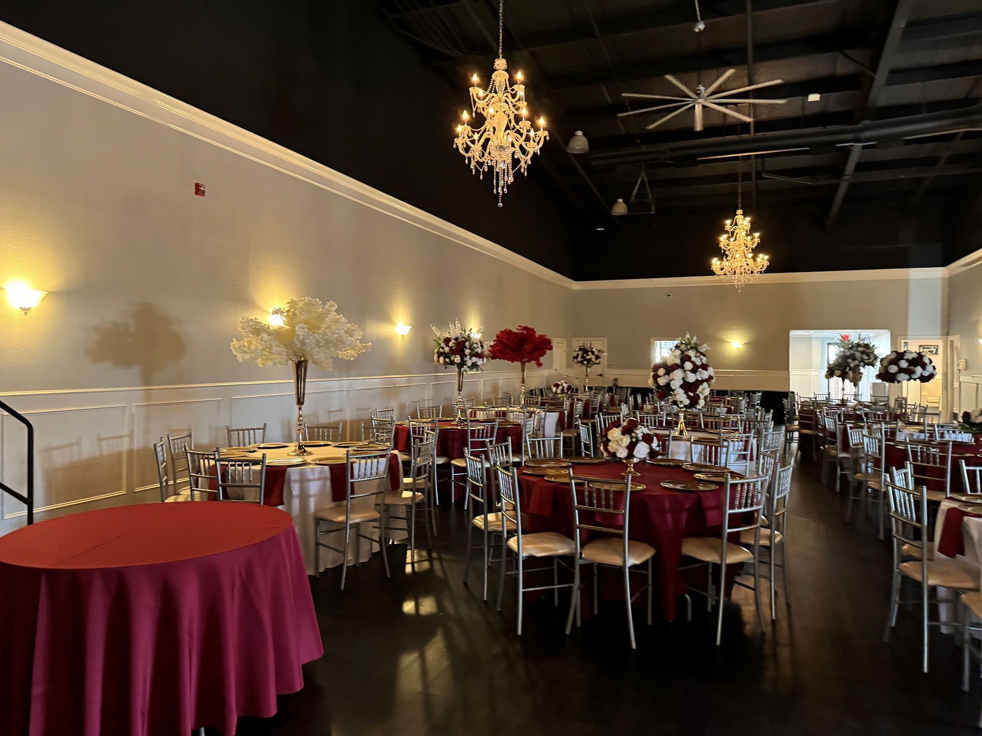 Elegant banquet hall with round tables, gold chairs, chandeliers, and red table linens in a spacious room