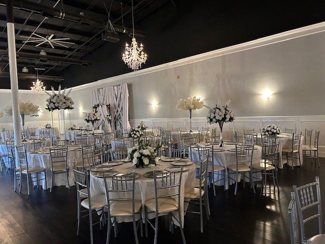 Elegant banquet hall with round tables, white chairs, floral centerpieces, and chandeliers under soft lighting