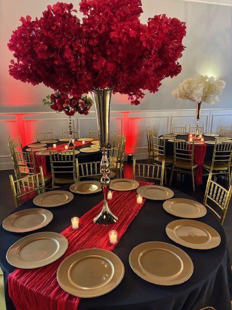 Elegant banquet table with gold plates, a red runner, and tall red floral centerpiece in a decorated hall