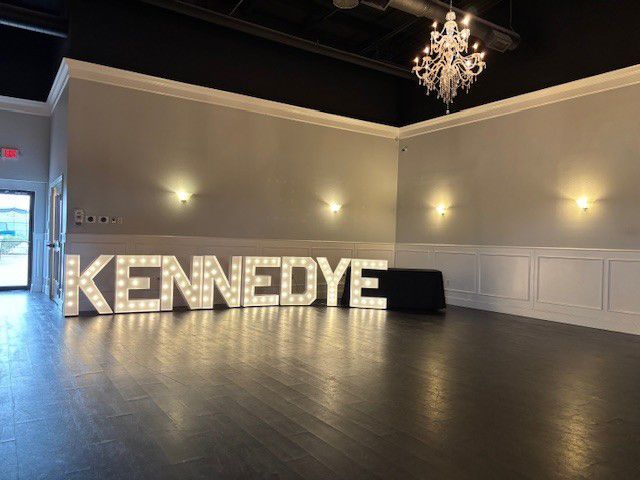 Illuminated “KENNEDYE” marquee letters in a dim event hall with chandeliers and gray walls