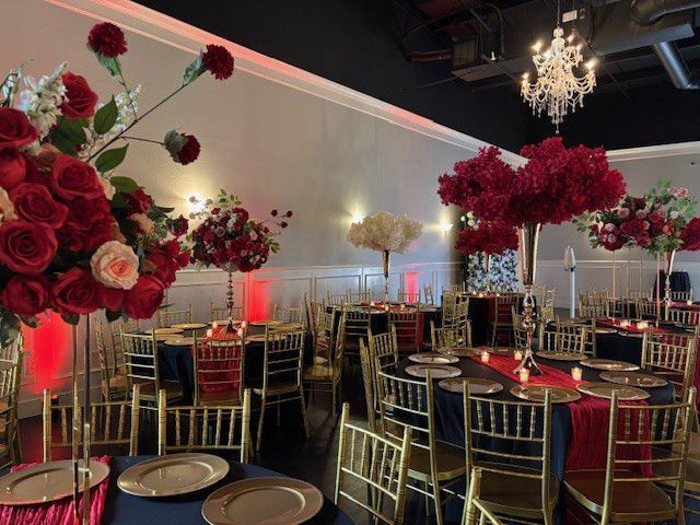 Elegant banquet hall with gold chairs, black tablecloths, and red floral centerpieces under a chandelier.