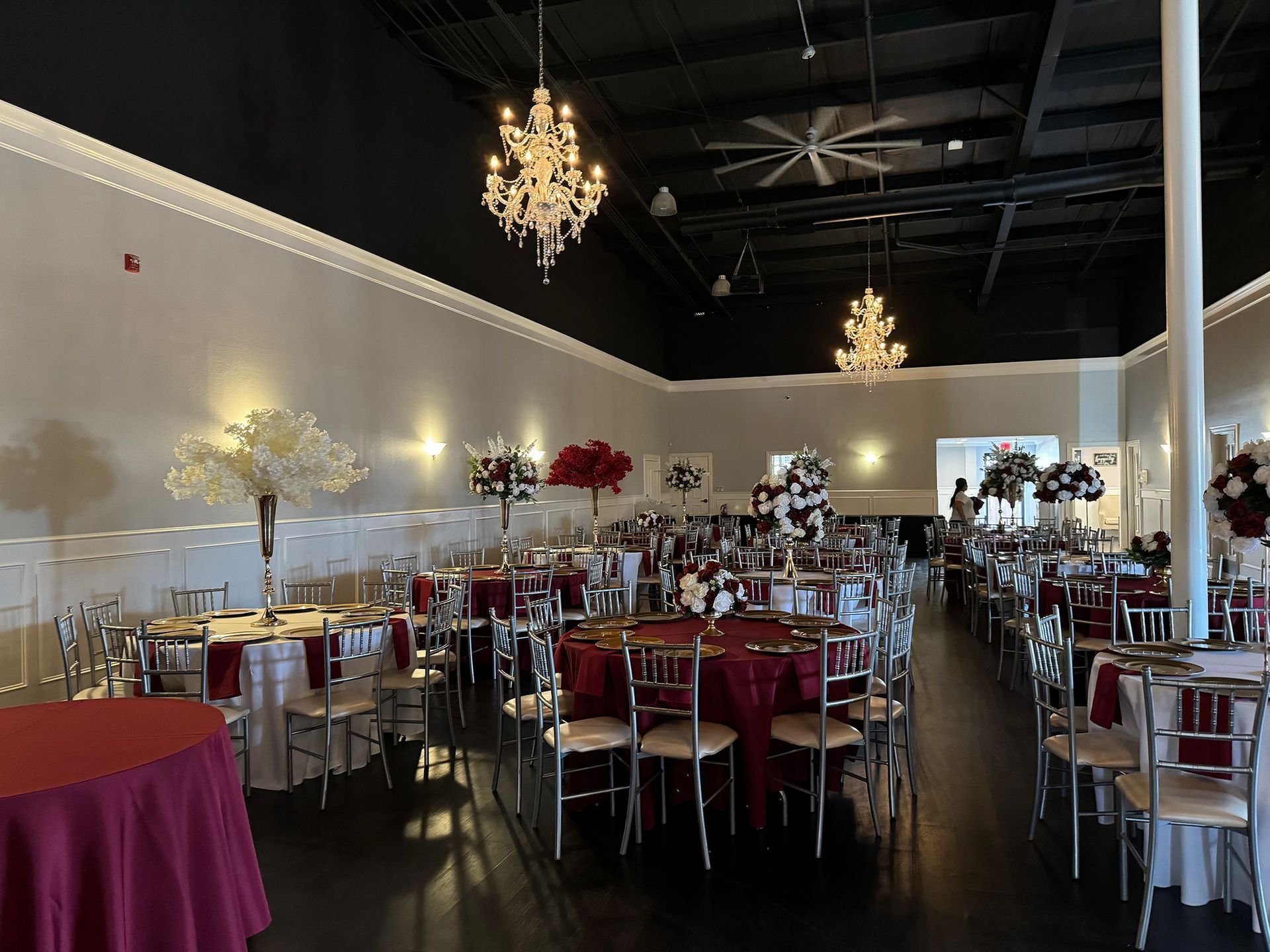 Elegant banquet hall with round tables, silver chairs, burgundy tablecloths, and chandeliers overhead
